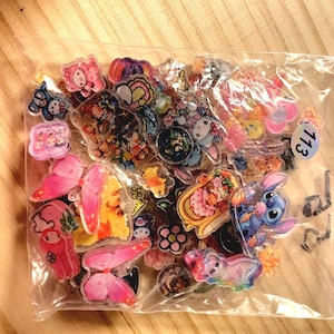 May include: A clear plastic bag filled with a variety of colorful acrylic charms featuring cartoon characters, butterflies, and flowers. A small white circular sticker with the number 113 is attached to the exterior of the plastic packaging.