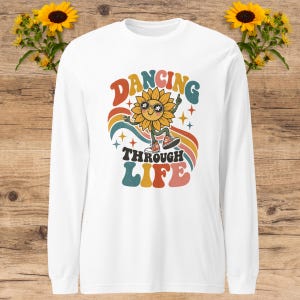 May include: A white long-sleeve shirt featuring a graphic of a smiling sunflower wearing sunglasses and trainers. The text DANCING THROUGH LIFE is printed in colourful, retro-style lettering, surrounded by a rainbow arc and small star shapes.