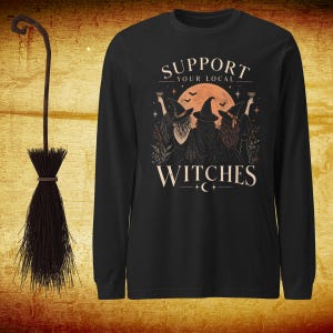 May include: A black long-sleeve shirt featuring a graphic of three witches in hats standing before a large orange moon. The text reads SUPPORT YOUR LOCAL WITCHES. Beside the shirt is a dark brown broom with a long handle and bristled end.