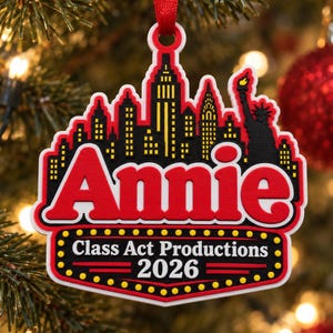 Puede incluir: Un adorno decorativo que presenta la silueta del horizonte de la ciudad de Nueva York con la Estatua de la Libertad en negro y amarillo. La palabra Annie está impresa en letras rojas grandes sobre un letrero estilo marquesina Class Act Productions 2026.