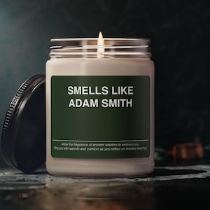 May include: A lit candle in a clear glass jar with a dark green label featuring the white text "SMELLS LIKE ADAM SMITH" and smaller text below. The jar has a black metal lid resting to the left side, and the candle wax is a light cream colour.
