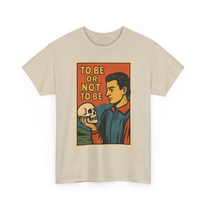 Könnte beinhalten: Ein beiges kurzärmeliges T-Shirt mit einem Grafikdruck einer Person, die einen Schädel betrachtet. Der Text TO BE OR NOT TO BE ist in gelben Blockbuchstaben gedruckt. Die Illustration verwendet eine Farbpalette aus Orange, Blau, Grün und Gelb.