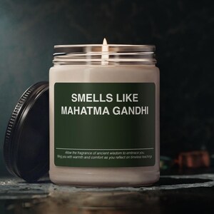 May include: A clear glass candle jar with a lit wick and a black lid placed to the side. The label is dark green with white text that reads: SMELLS LIKE MAHATMA GANDHI, followed by smaller text: Allow the fragrance of ancient wisdom to embrace you.