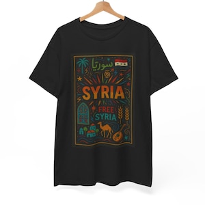 Puede incluir: Una camiseta negra de manga corta con un estampado gráfico rectangular que incluye el texto SYRIA y FREE SYRIA. El diseño presenta ilustraciones coloridas de una palmera, la bandera siria, un camello, un instrumento musical y edificios.