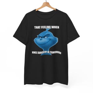 Op de afbeelding: Een zwart t-shirt met korte mouwen en een afbeelding van een blauw, ondeugend uitziend personage met een grijns. Boven het personage staat de tekst "THAT FEELING WHEN" en onder het personage staat "KNEE SURGERY IS TOMORROW".