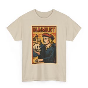 Könnte beinhalten: Ein beiges Kurzarm-T-Shirt mit einem Grafikdruck einer Person mit roter Mütze, die einen Totenkopf hält. Der Text HAMLET ist in Rot oben auf der rechteckigen Illustration gedruckt, die zudem eine steinerne Burg im Hintergrund zeigt.