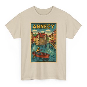 Puede incluir: Una camiseta beige de manga corta con un estampado gráfico rectangular de una escena de canal en Annecy. La ilustración muestra a una persona en un pequeño bote rojo, un edificio de piedra, casas coloridas y la palabra ANNECY en letras amarillas.