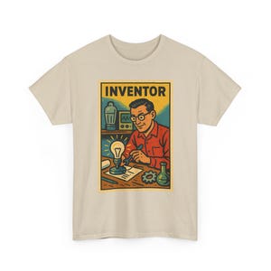 Peut inclure: Un t-shirt beige à manches courtes avec une impression graphique représentant une personne à lunettes en chemise rouge, travaillant à un bureau avec une ampoule allumée, une clé, un engrenage et une fiole. Le mot INVENTOR est écrit en noir.