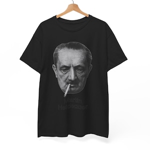 Könnte beinhalten: Ein schwarzes Kurzarm-T-Shirt mit einem graustufigen Fotodruck eines Mannes mit Schnurrbart, der eine Zigarette raucht. Unter dem Gesicht ist der Text Martin Heidegger in einer dunklen, dezenten Schrift auf der Brustmitte aufgedruckt.