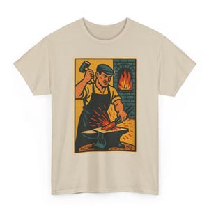以下が含まれることがあります： ベージュの半袖Tシャツ。帽子とエプロンを身につけた鍛冶屋が、ハンマーで金床の上の赤く光る金属を叩いている様子を描いたグラフィックイラストが中央にあり、背景にはレンガ造りの炉とオレンジ色の炎が描かれています。