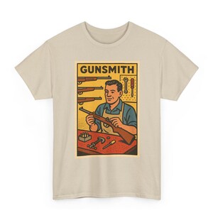 Può includere: Una t-shirt beige con un'illustrazione grafica di un armaiolo al lavoro su un banco da lavoro. La stampa mostra una persona con un grembiule che tiene in mano un fucile, con attrezzi su un pannello, altri fucili alla parete e la scritta GUNSMITH.