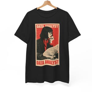 Pode incluir: Uma t-shirt preta de manga curta com uma impressão gráfica retangular. A estampa mostra uma pessoa de cabelo escuro e óculos, vestindo uma camisa vermelha e um blazer escuro, lendo um documento. O texto DATA ANALYST aparece em vermelho.