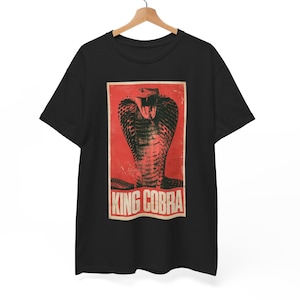 Puede incluir: Una camiseta negra de manga corta con un estampado gráfico rectangular en el pecho. El estampado muestra un fondo rojo con una ilustración detallada de una cobra real con la capucha desplegada y la boca abierta. El texto KING COBRA está en blanco.