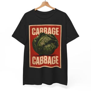 May include: A black short-sleeved t-shirt featuring a rectangular graphic print. The design displays a dark green head of cabbage centred on a red background, with the word CABBAGE printed in bold, off-white capital letters above and below it.