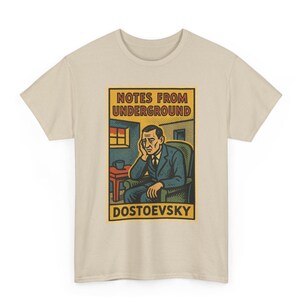 May include: A beige short-sleeve t-shirt featuring a graphic print of a person sitting in a chair with the text "NOTES FROM UNDERGROUND" and "DOSTOEVSKY" in a rectangular frame. The illustration uses muted tones of blue, yellow, and red.