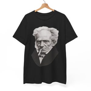 May include: A black short-sleeve t-shirt featuring a large, detailed white graphic print of the philosopher Arthur Schopenhauer. The portrait shows him with his signature wild hair, a dark coat, and a cigarette held between his lips.