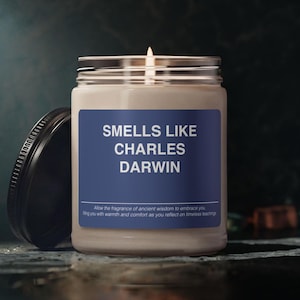 May include: A clear glass jar candle with a lit wick and a black lid placed to the side. The label is blue with white text that reads SMELLS LIKE CHARLES DARWIN, followed by smaller text that says Allow the fragrance of ancient wisdom to embrace you.