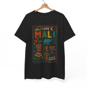 May include: A black short-sleeve t-shirt featuring a colorful graphic print with the text I LOVE MALI and LAND OF BAOBABS. The design includes illustrations of a person, an elephant, drums, a baobab tree, a heart, and a crescent moon.