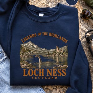 May include: A navy blue crewneck sweatshirt featuring a graphic print of a lake with a long-necked creature, mountains, and a stone castle. The text reads "LEGENDS OF THE HIGHLANDS" at the top and "LOCH NESS SCOTLAND" at the bottom in orange.