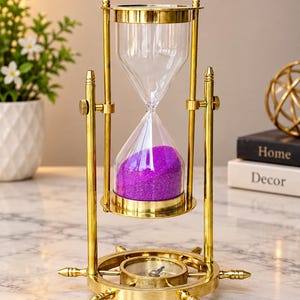 Könnte beinhalten: Eine Sanduhr aus Messing mit drehbarem Rahmen, einem Sockel in Form eines Steuerrads und einem integrierten Kompass. Die Glaskolben enthalten leuchtend violetten Sand und die Metallstruktur hat ein poliertes, goldfarbenes Finish.