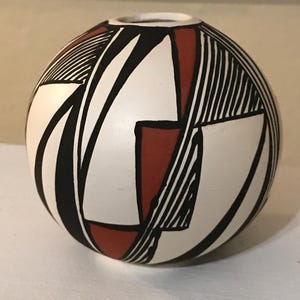May include: A spherical ceramic vase featuring a matte white base decorated with bold black geometric lines and solid terracotta red shapes. The design consists of intersecting triangles and parallel line patterns covering the rounded surface.