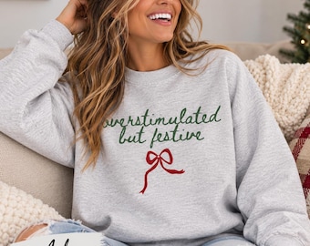 May include: A light gray crewneck sweatshirt featuring the dark green text "overstimulated but festive" written in a cursive script, positioned above a red graphic illustration of a tied ribbon bow. The fabric has a heathered texture.
