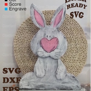 Könnte beinhalten: Eine bemalte Holzhasenfigur mit einer rosa Herz-Nase und rosa Innenohren. Die Figur ist grau mit weißen Pinselstrichen und sitzt auf einem Sockel. Der Text auf dem Bild lautet "LASER READY SVG".