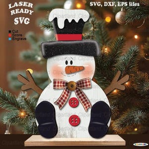 May include: A wooden snowman cutout with a black top hat, red and white checkered bow, and a carrot nose. The snowman is sitting on a wooden shelf. The image includes the text "LASER READY SVG, DXF, EPS files".