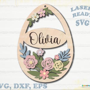 May include: Wooden Easter egg-shaped decoration with the name "Olivia" in script. The egg is adorned with pastel-colored flowers and leaves. The text "LASER READY SVG" and "SVG, DXF, EPS" are also present.