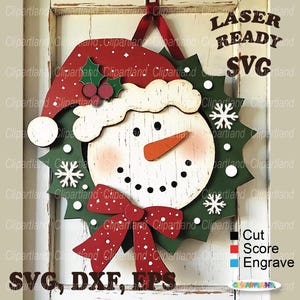May include: A wooden snowman wreath with a red Santa hat, bow, and holly accents. The wreath has a green base with white snowflakes and a snowman face. Text on the image reads "LASER READY SVG" and "SVG, DXF, EPS".