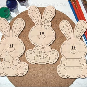 May include: Three unfinished wooden bunny cutouts with black outlines, ready for painting. One bunny holds an egg, another has a flower, and the third is plain. Paint pots and brushes are in the background, suggesting a craft project.