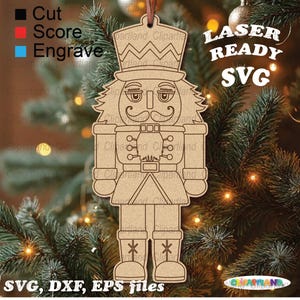May include: A wooden laser-cut Christmas ornament shaped like a nutcracker soldier. The ornament features a detailed design of a hat, mustache, and uniform. The text "LASER READY SVG" and "SVG, DXF, EPS files" are visible. It hangs on a Christmas tree.