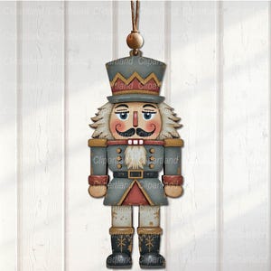 May include: A wooden Christmas ornament shaped like a nutcracker soldier. The ornament features a gray coat, a red and gold trim, and a black hat. The nutcracker has a white beard and mustache, and is hanging from a brown string.