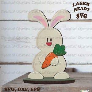 May include: A white bunny rabbit holding a carrot with green leaves. The bunny is standing on a brown base. The image is labeled "Laser Ready SVG, DXF, EPS".
