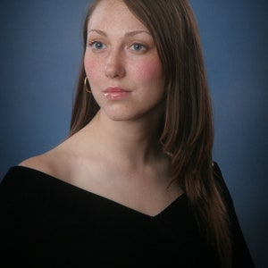 May include: A portrait of a person with fair skin, freckles, and long brown hair. They are wearing a black off-the-shoulder top and gold hoop earrings. The background is a gradient of blue.