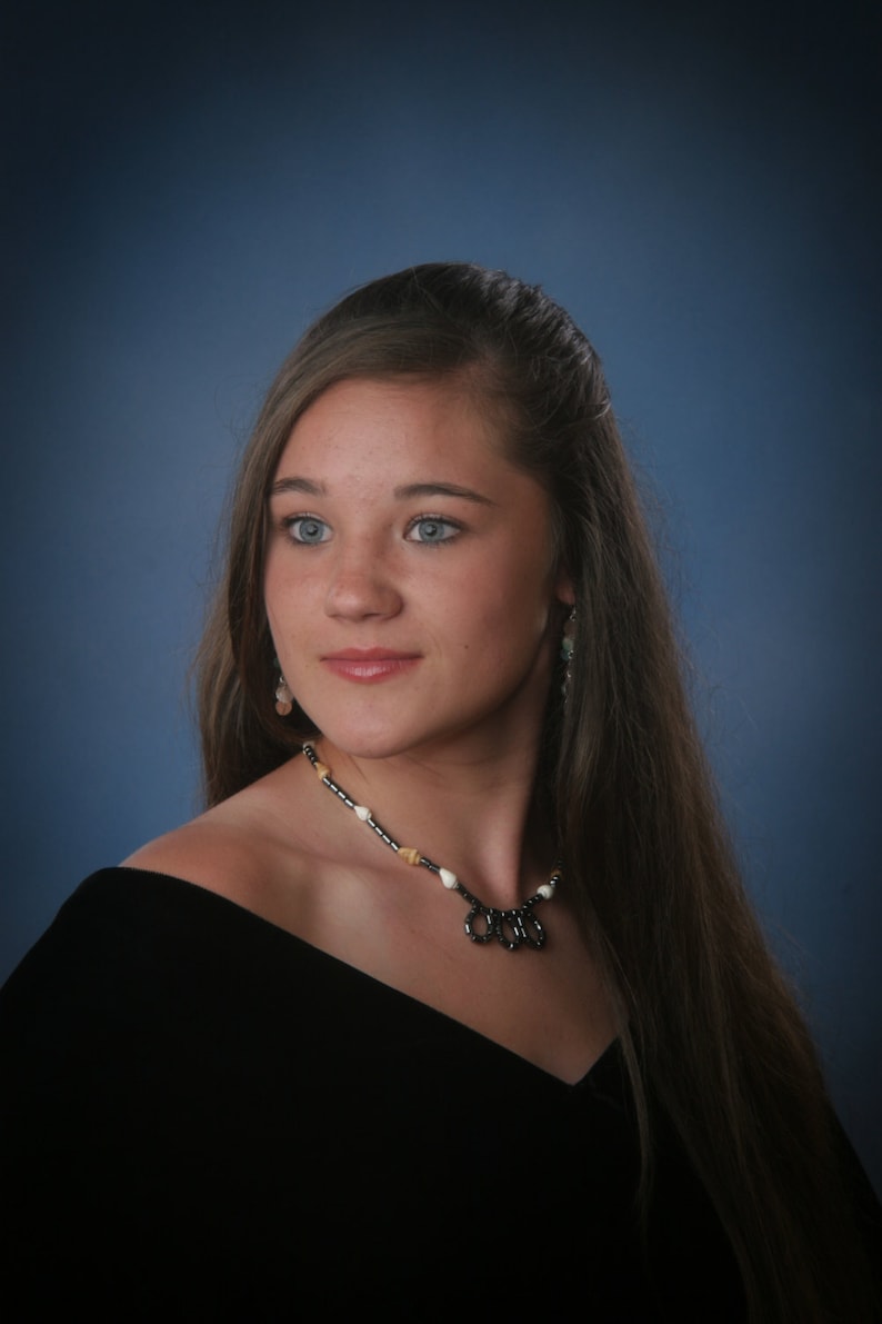 May include: A portrait of a person with long brown hair, blue eyes, and a black off-the-shoulder top. They are wearing a necklace with black, white, and tan beads. The background is a gradient of blue.
