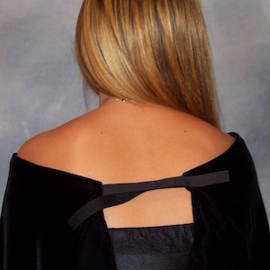 May include: A black velvet cape with a satin panel in the back. The cape has a low neckline and a black ribbon detail at the back. The person's blonde hair is visible.