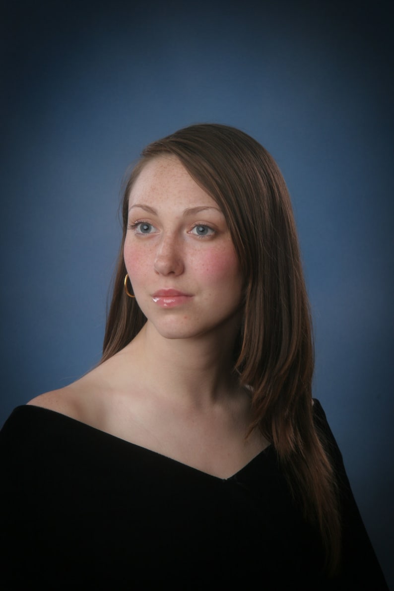 May include: A portrait of a person with long brown hair, blue eyes, and freckles. They are wearing a black off-the-shoulder top and gold hoop earrings. The background is a gradient of blue.