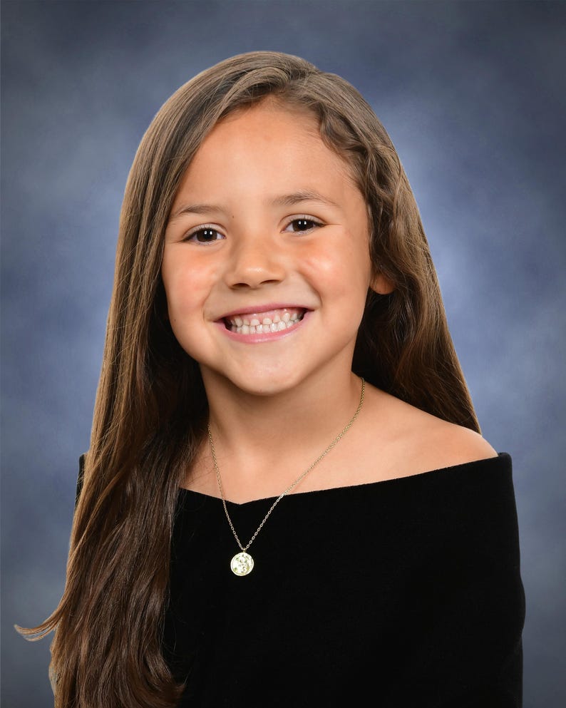 Puede incluir: Retrato de una ni&ntilde;a sonriente con cabello casta&ntilde;o largo, vistiendo un top negro con hombros descubiertos y un collar con colgante dorado. El fondo es un degradado de azul y gris.