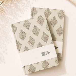 Pode incluir: Dois cadernos de capa dura com uma capa bege clara e um padrão floral repetitivo em forma de diamante em tons de castanho, azul e verde. Cada caderno está envolvido por uma faixa de papel branca com o texto ruled handmade papers.