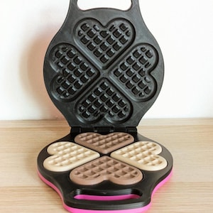 Peut inclure: Un gaufrier noir avec une base rose et un couvercle articulé. L'intérieur présente quatre moules en forme de cœur avec un motif en grille. À l'intérieur des moules se trouvent quatre gaufres en forme de cœur de couleurs beige et marron.
