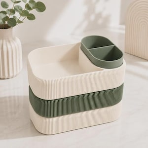 Può includere: Un organizer da scrivania impilabile con una base color crema e una sezione centrale verde scuro, entrambe con una texture a coste verticali. Il vassoio superiore è color crema con un inserto verde scuro diviso in due scomparti separati.