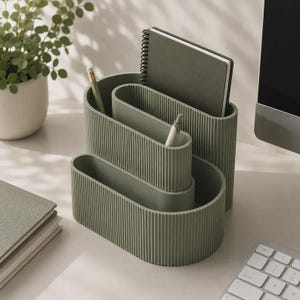 Pode incluir: Um organizador de secretária verde-sálvia com um design oval e canelado, apresentando três compartimentos escalonados de alturas variadas. O organizador contém um caderno de espiral e duas canetas, oferecendo uma solução de arrumação.