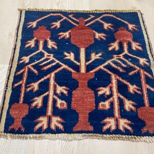 May include: A rectangular woven rug featuring a deep blue background with a central terracotta-colored vase motif. The design includes stylized branches and pomegranate-like shapes in terracotta and cream, finished with short fringe on the edges.