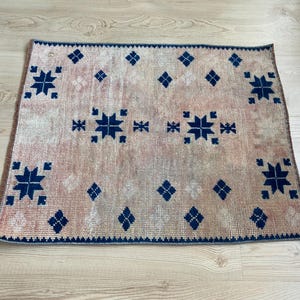 May include: A rectangular rug featuring a faded peach and beige background with a repeating pattern of dark navy blue geometric star motifs and small diamond shapes. The rug has a dark navy blue border along all four edges of the fabric.