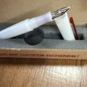 May include: A white fountain pen with a silver-toned metal nib and a translucent white cap featuring a dark red clip. The pen is resting in a gray foam-lined cardboard box that has the text "OF SATISFIED CUSTOMERS" printed on the front edge.