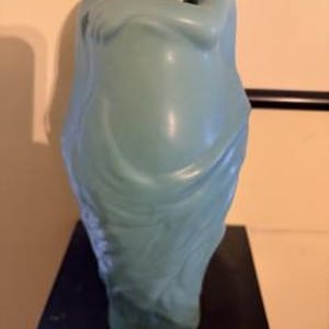 May include: A matte, light blue ceramic sculpture depicting a figure in a curled, seated position with arms wrapped around the knees. The surface features smooth, flowing contours and draped lines that create a sense of movement and form.