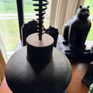 May include: A dark, textured, spherical metal object with a flat top and a vertical, spiralling metal coil extending upwards from the centre. The object has a matte, weathered finish and is positioned on a dark, circular base with visible rivets.