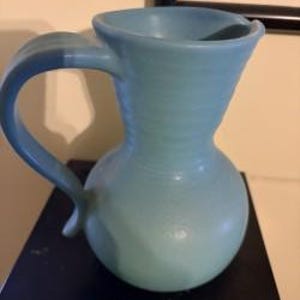 Może przedstawiać: Jasnoniebieski dzbanek ceramiczny z zaokrągloną podstawą, wąską szyjką i szerokim, rozszerzającym się brzegiem. Dzbanek posiada zakrzywiony uchwyt przymocowany z boku, z widocznymi poziomymi prążkami wzdłuż szyjki oraz gładkim, matowym wykończeniem.
