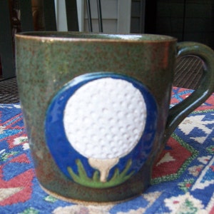 May include: A green ceramic mug with a white golf ball design. The golf ball is surrounded by a blue circle and green grass.